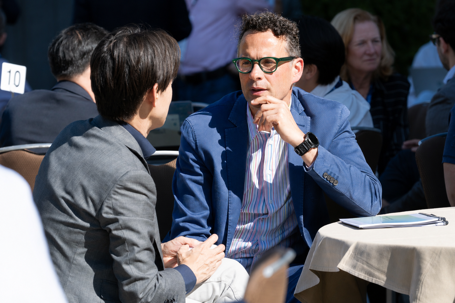 Two men are engaged in a conversation at an outdoor event, with other people seated at tables in the background.