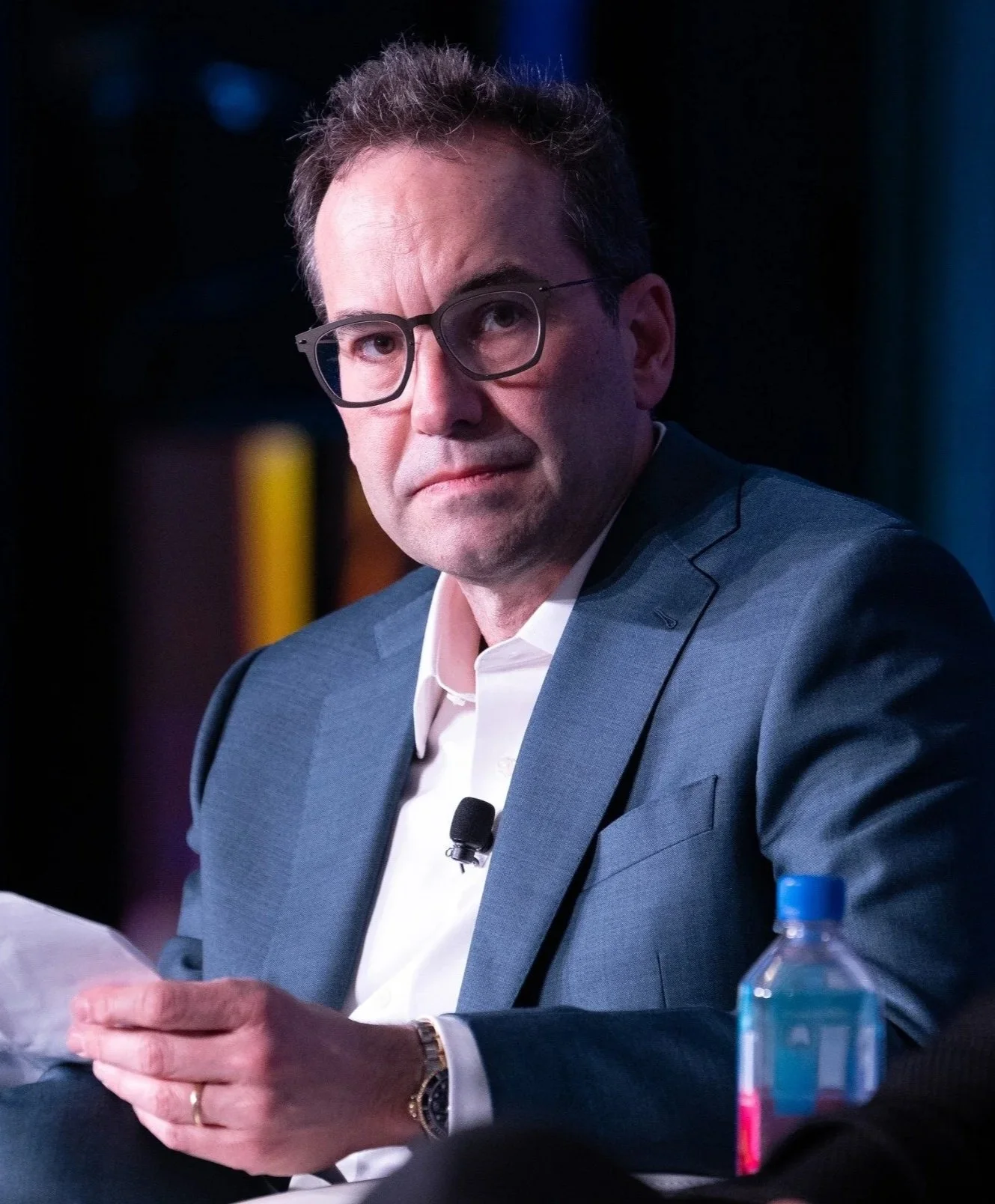 A middle-aged man with glasses wearing a blue suit and white shirt, sitting at a table with a water bottle and papers, looking to the side in a professional setting.