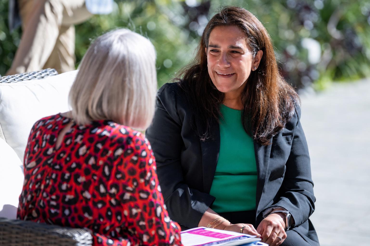 Two women sitting outdoors, engaged in conversation, with trees in the background. One woman has gray hair and wears a red patterned top, while the other has long dark hair, wears a green shirt and dark blazer.