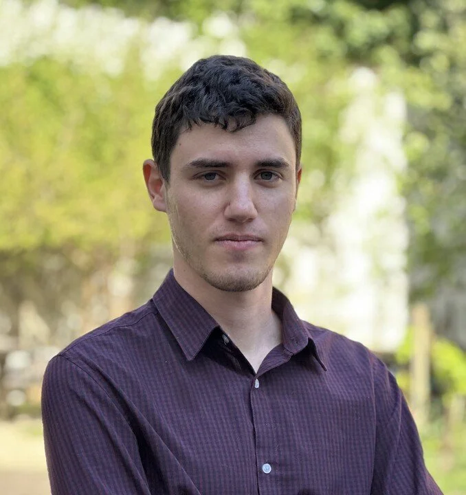 A young man in a purple shirt stands looking at the camera, with head, shoulders, and chest visible in the picture. The background is blurred, but appears to be some sort of vegetation.