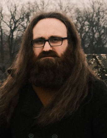 A man with long hair and glasses looks at the camera. The picture shows head, shoulders, and upper chest against a white background.