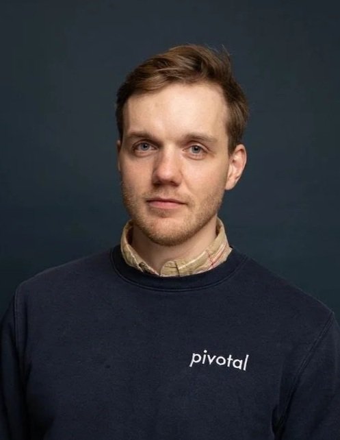 Portrait of a young man with light skin, brown hair, and a slight smile, wearing a black polo shirt outdoors, with a blurred background of a building and greenery.