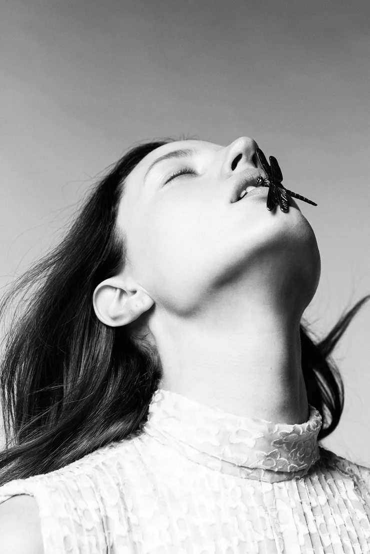 A black and white photo of a woman with long dark hair, her head tilted back with eyes closed, and a dragonfly perched on her lips.