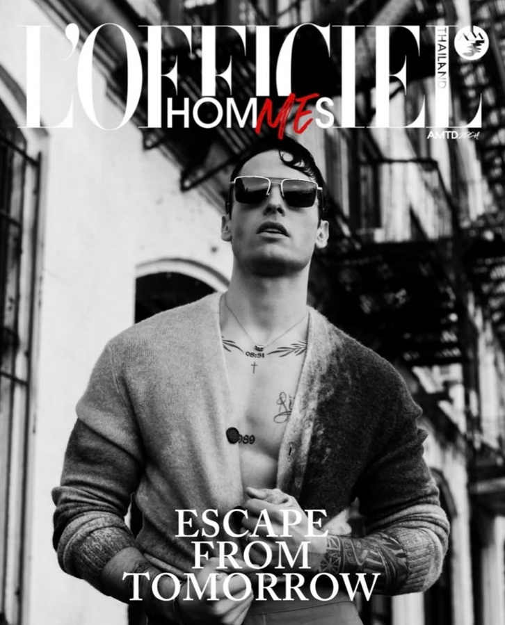 Black-and-white photo of a young man with tattoos, wearing sunglasses and an open cardigan, standing outdoors in an urban setting with fire escape stairs in the background. The magazine cover features the text "L'OFFICIEL HOMMES THAILAND" and the hea