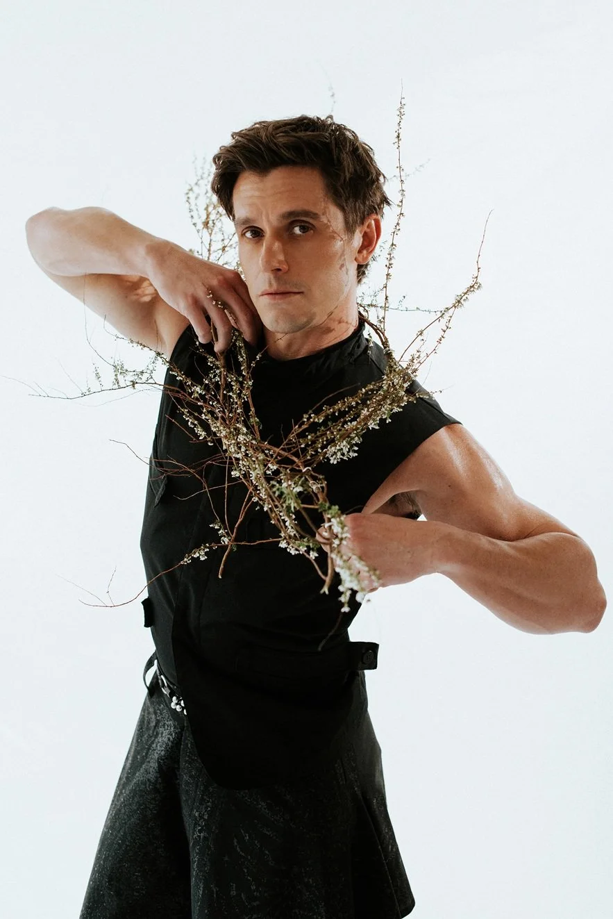 A man with short dark hair wearing a sleeveless black shirt and dark pants, posing with a serious expression while holding a branch of small white flowers and thin twigs in front of a plain white background.