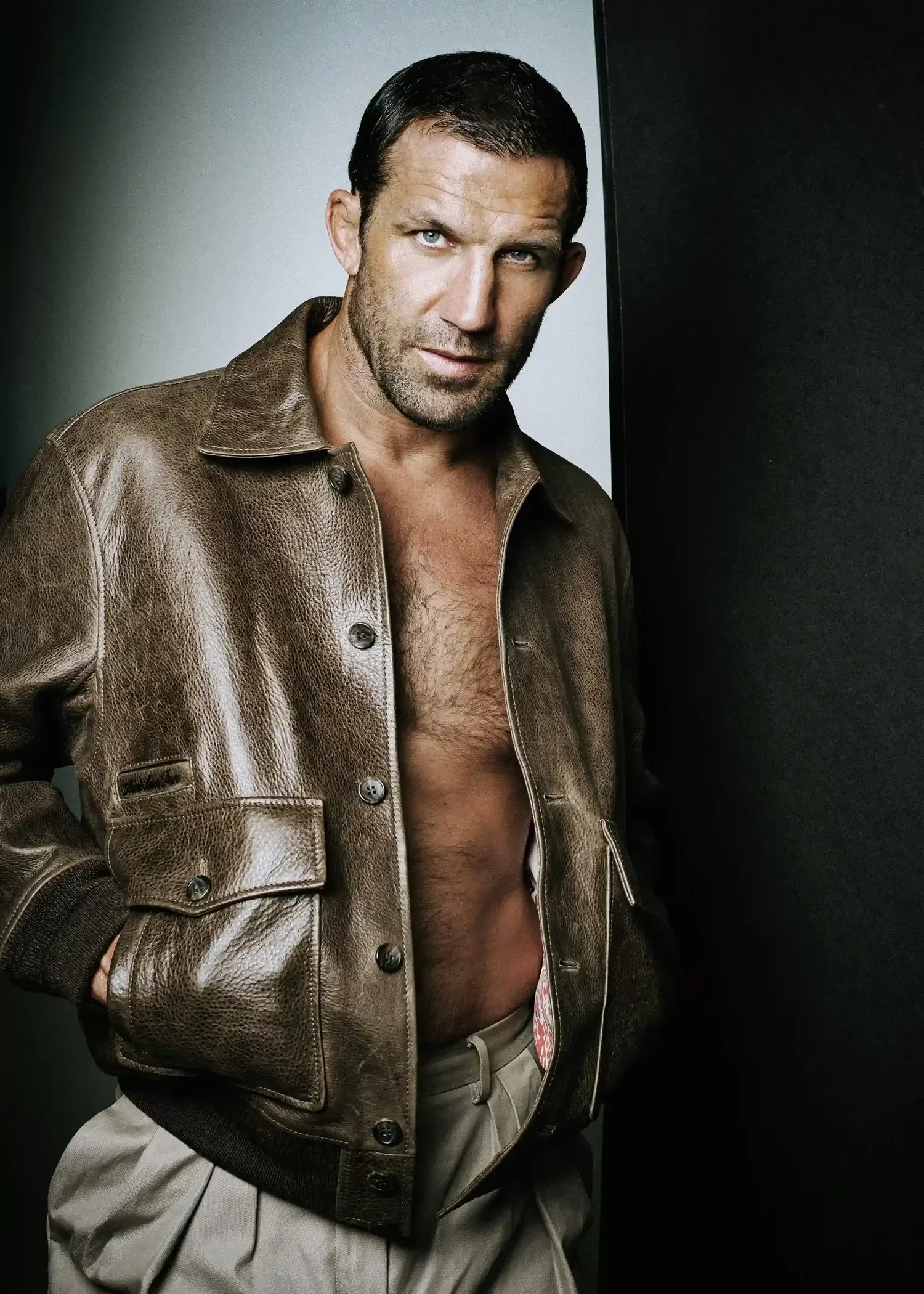 A man with dark hair and light eyes wearing a brown leather jacket with an open front, showing his bare chest, and beige pants against a plain gray and black background.