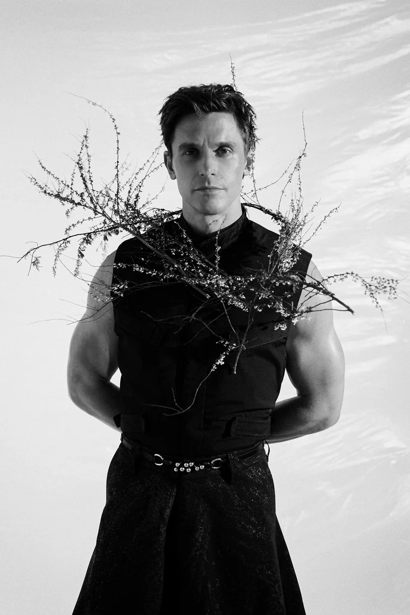 Black and white photo of a young man with dark hair, wearing a sleeveless black top, standing outdoors with a cloudy sky in the background. He has a serious expression and branches with small leaves or flowers crossing his chest.