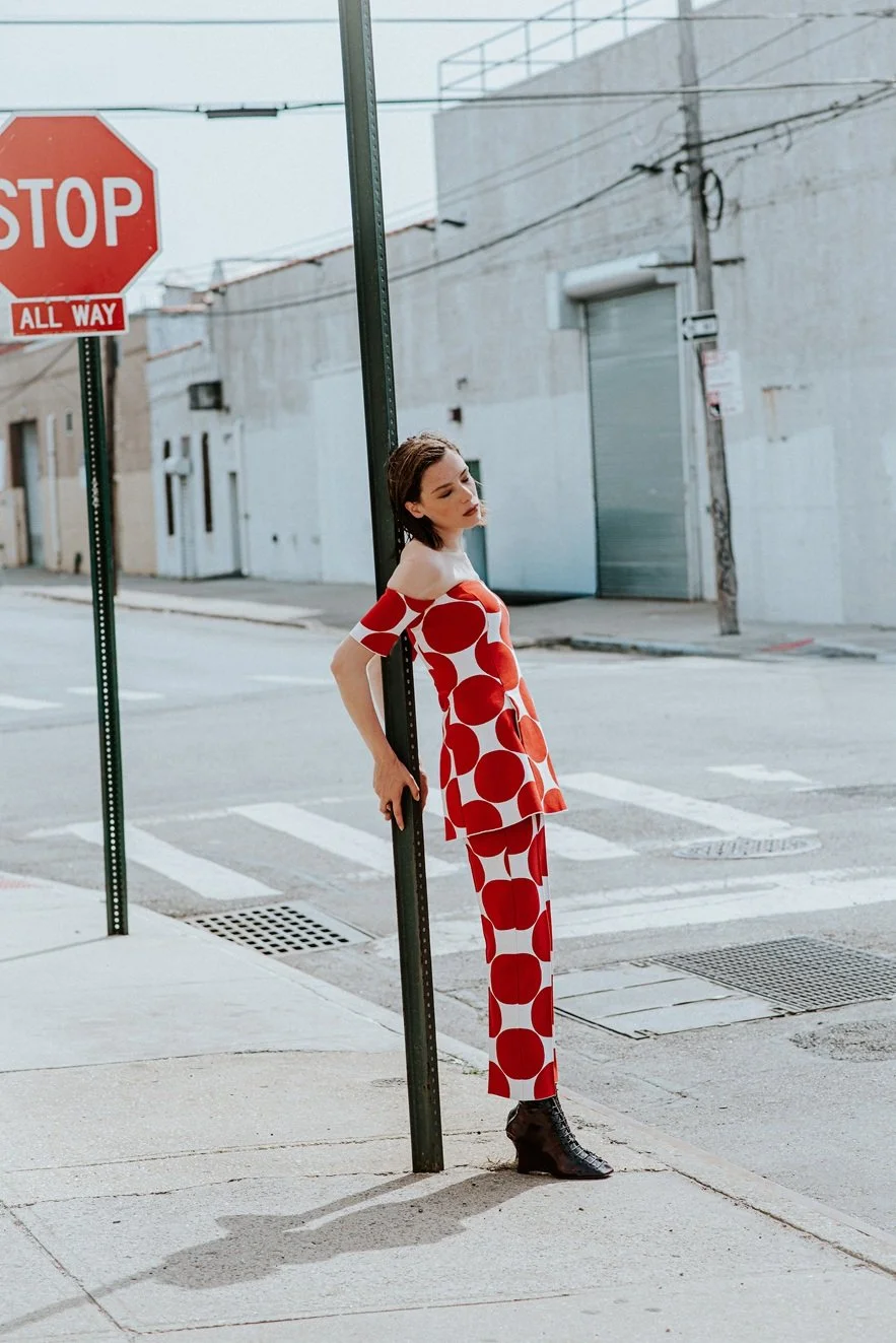 A woman in a red and white polka dot outfit leans against a pole at an empty street corner, with a stop sign visible in the upper left corner.