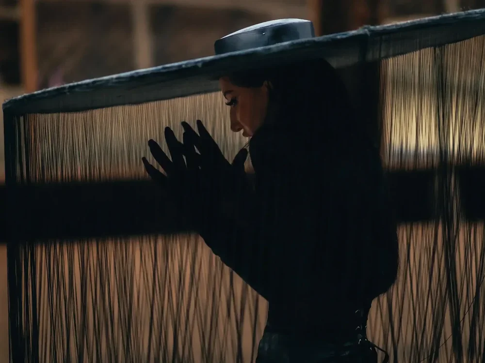 A woman in black gloves and a hat ties a black string or rope in a large, partially enclosed outdoor space with wooden beams and a bamboo or similar woven fence.