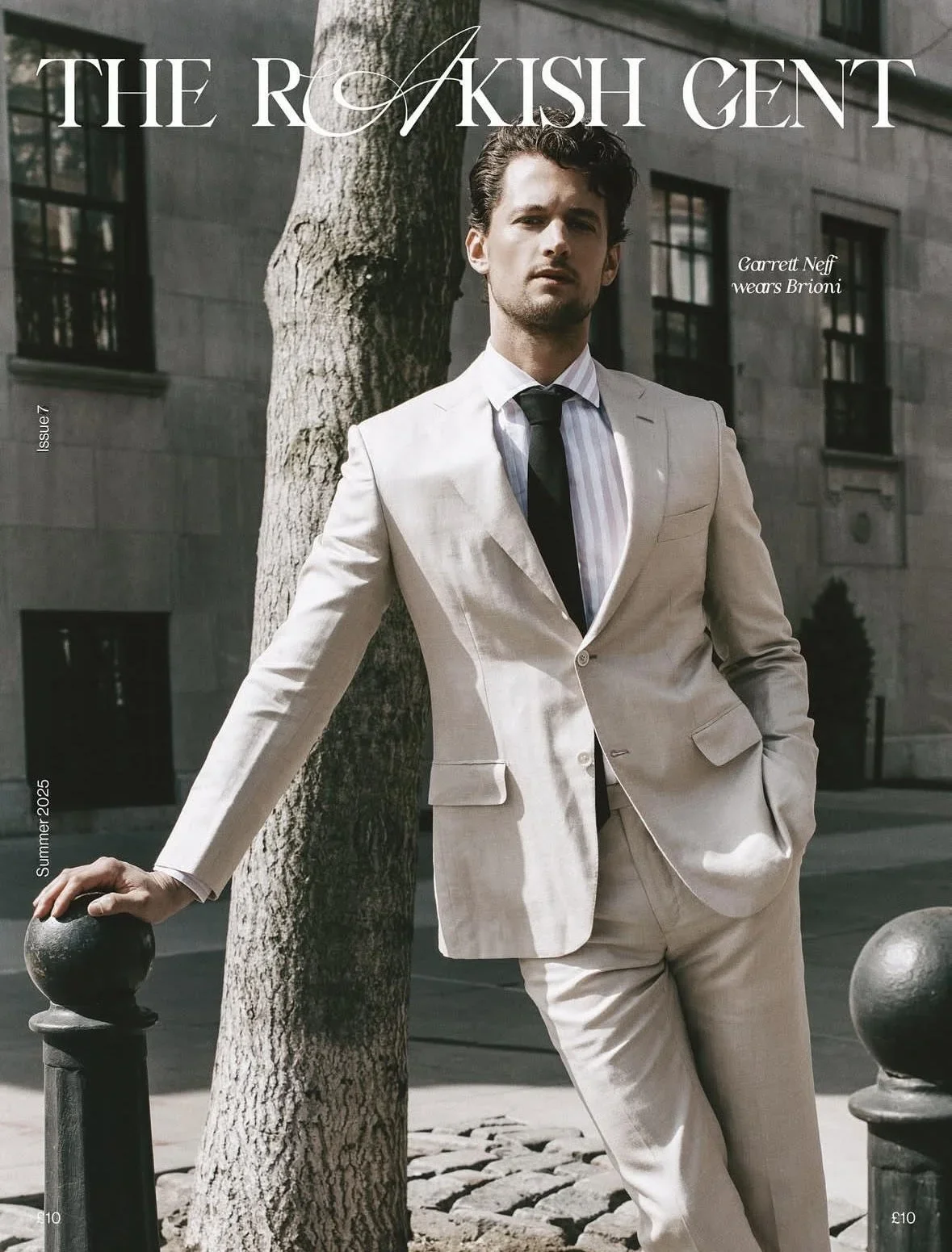 A man in a light-colored suit and striped shirt with a black tie, leaning against a black railing with one hand, standing next to a tree on a city street with gray stone buildings in the background.