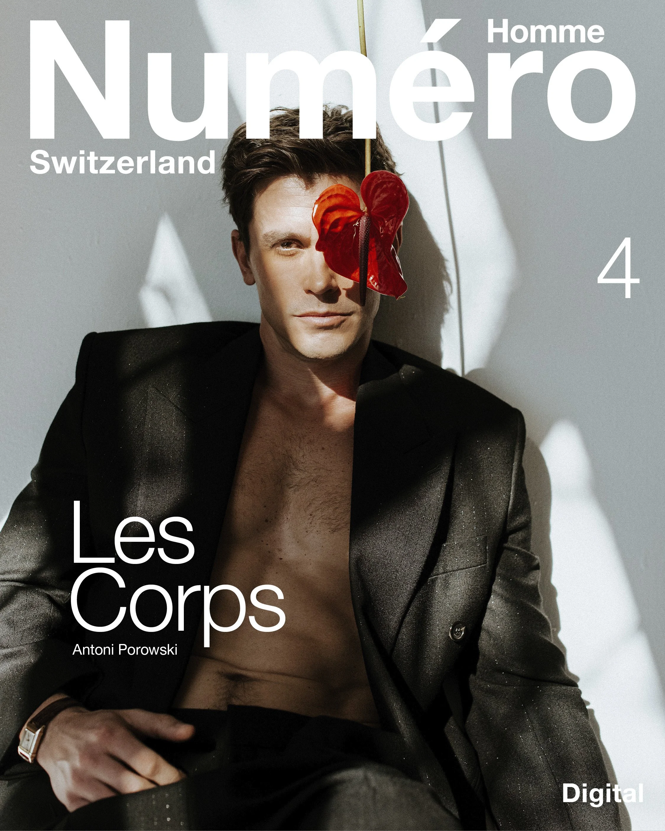 A man with brown hair, wearing a black blazer, partially shirtless, with a red and orange flower covering his right eye, sitting against a white wall with shadows.