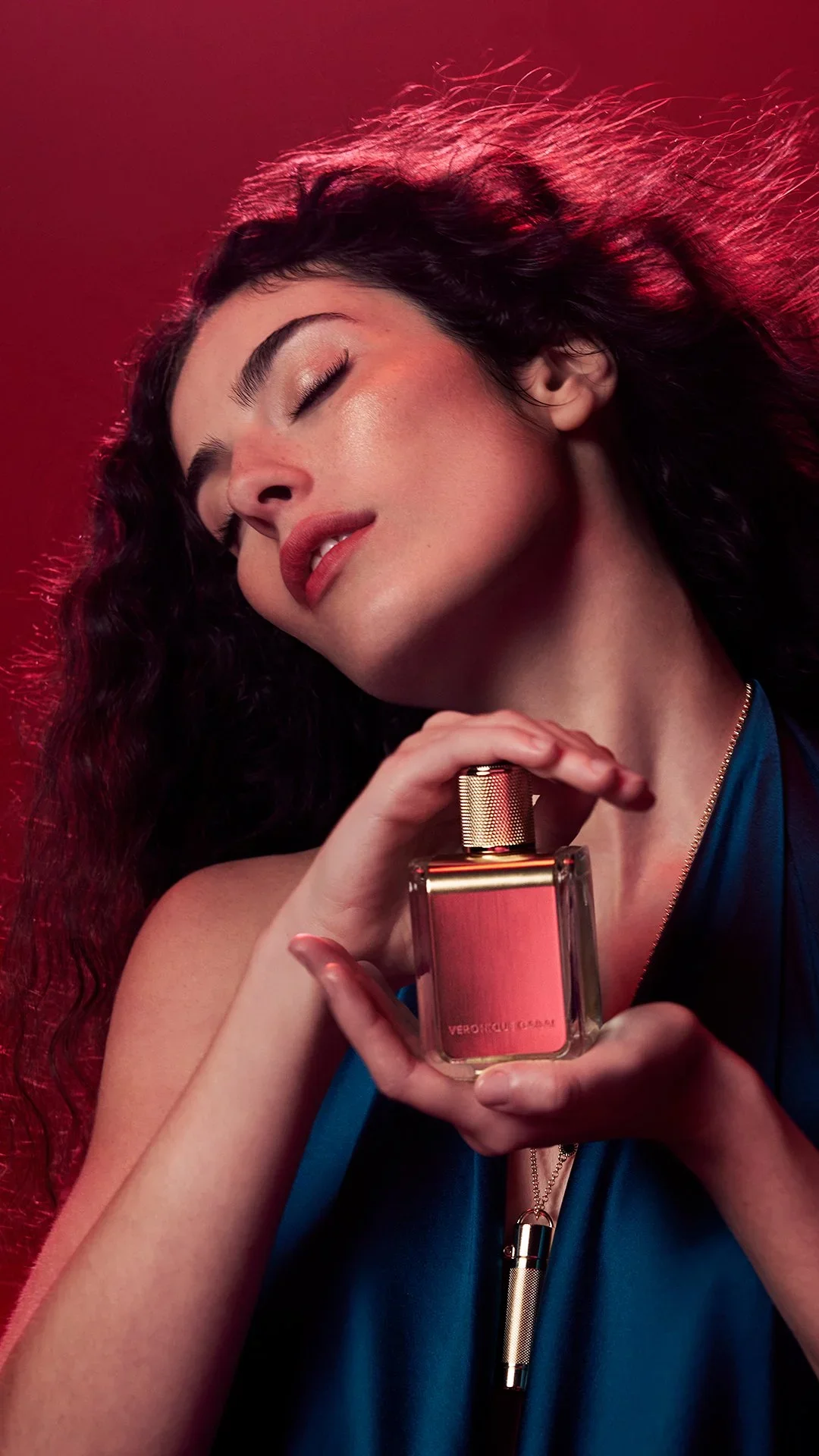 A woman with curly hair and closed eyes holding a perfume bottle labeled 'Vero Profumo GRAAM' against a red background.