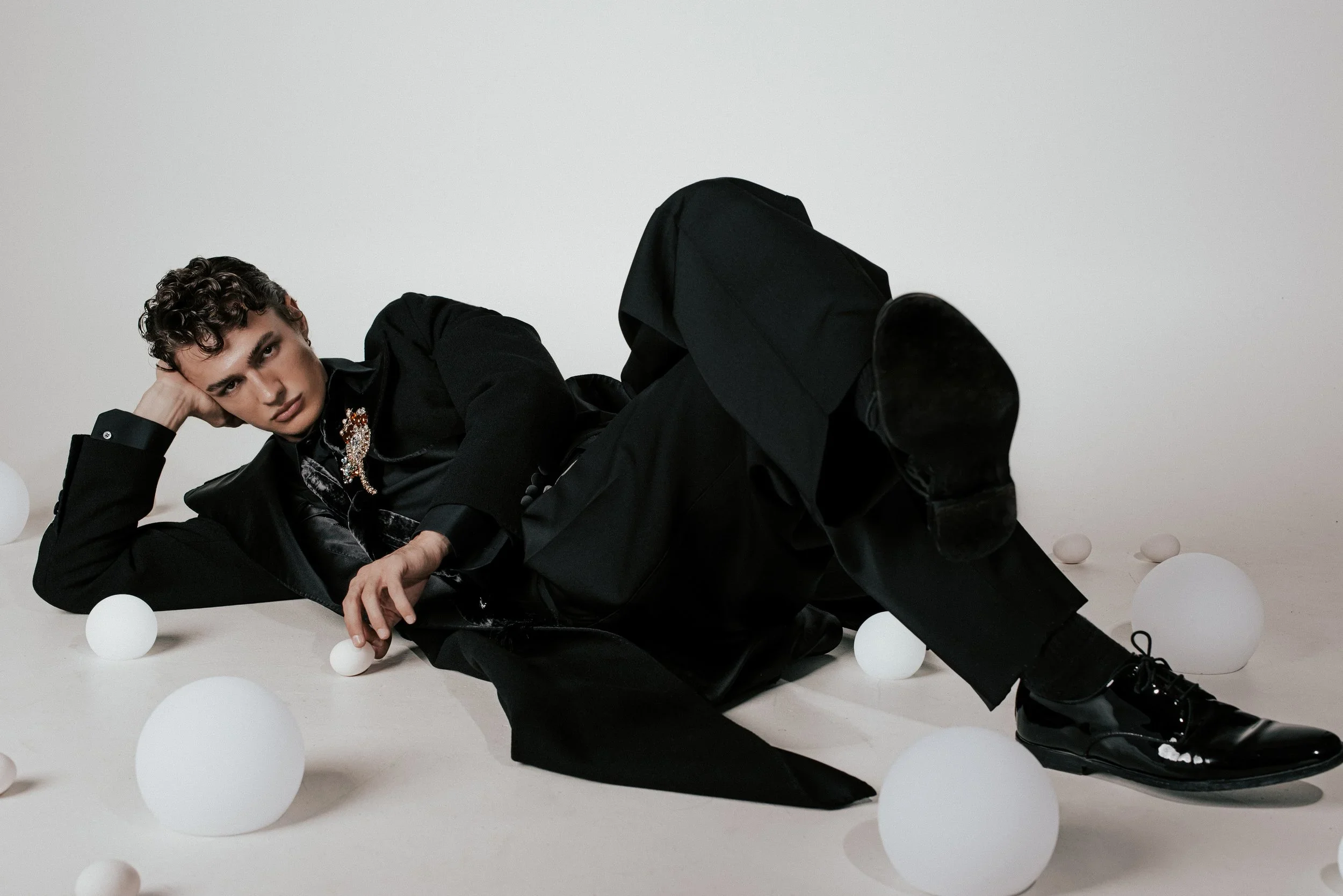 A young man with curly hair lying on the floor amidst white spherical objects, wearing a black outfit and looking at the camera.