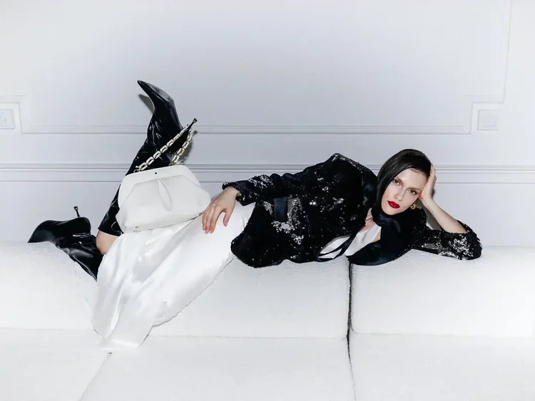 Woman lying on a white couch, dressed in a black sequin jacket, white skirt, and black boots, with a white purse, against a white wall.