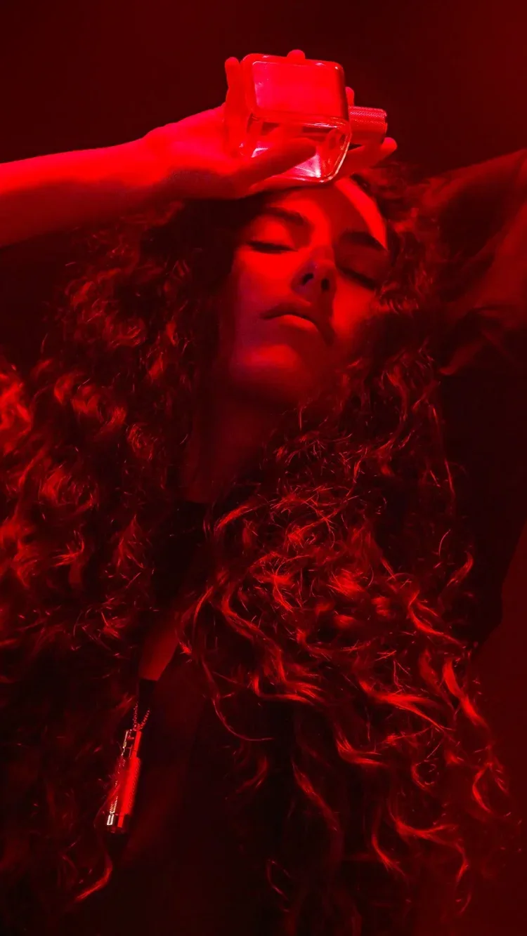 Woman with curly hair in red lighting, holding a small rectangular object above her forehead.