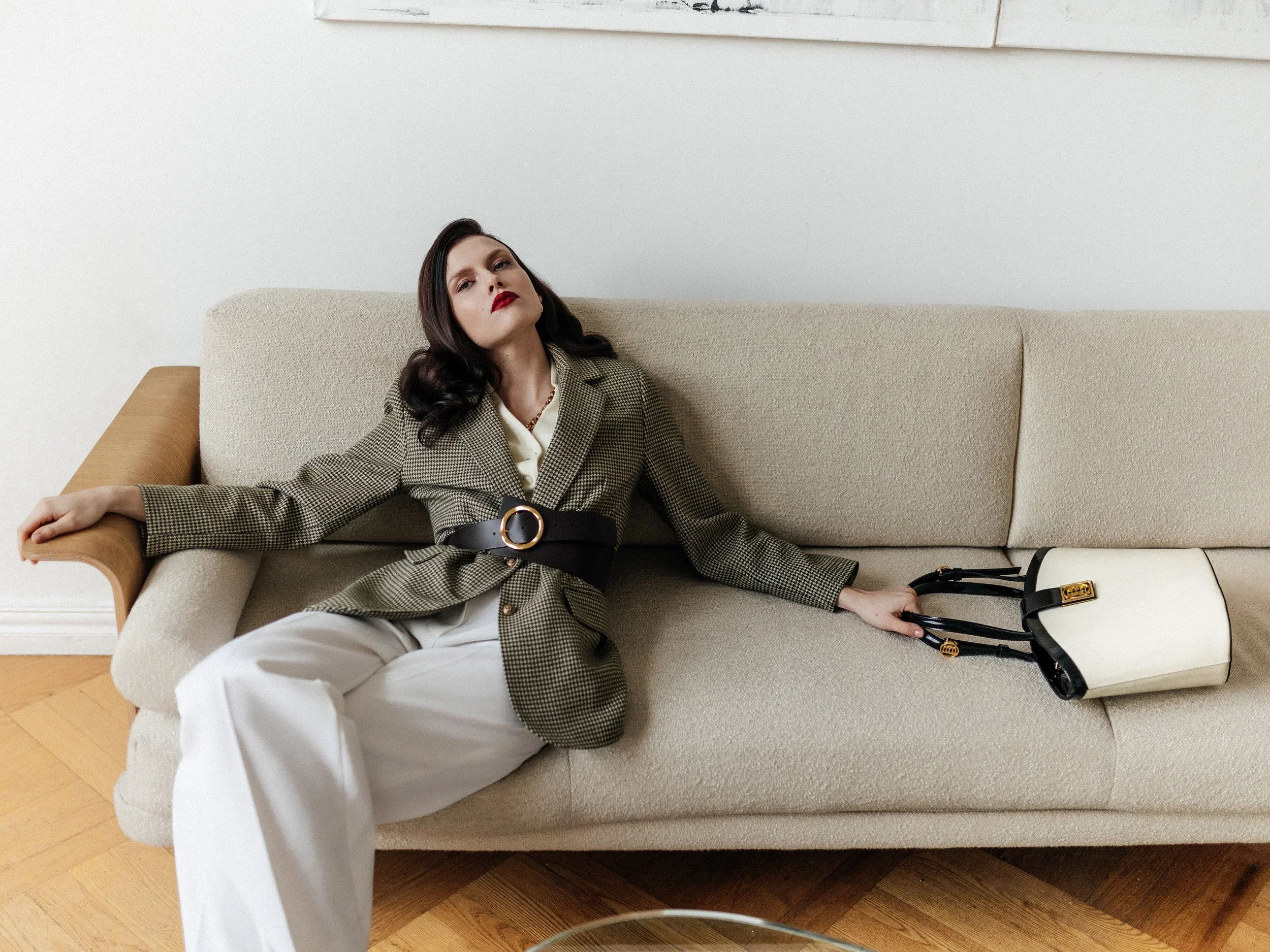 A woman with dark hair, red lipstick, wearing a beige checkered blazer and white pants, is lounging on a light beige sofa with her arm resting on the armrest, next to a beige and white handbag.