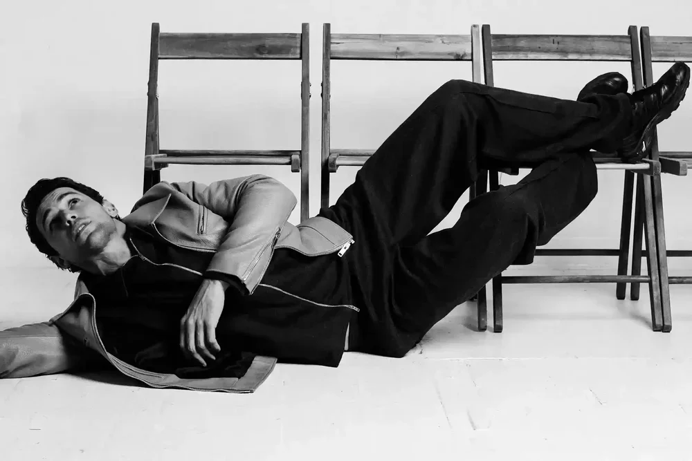A man lying on the floor next to a row of wooden chairs, looking up with his head on the floor and one arm extended, in black and white.
