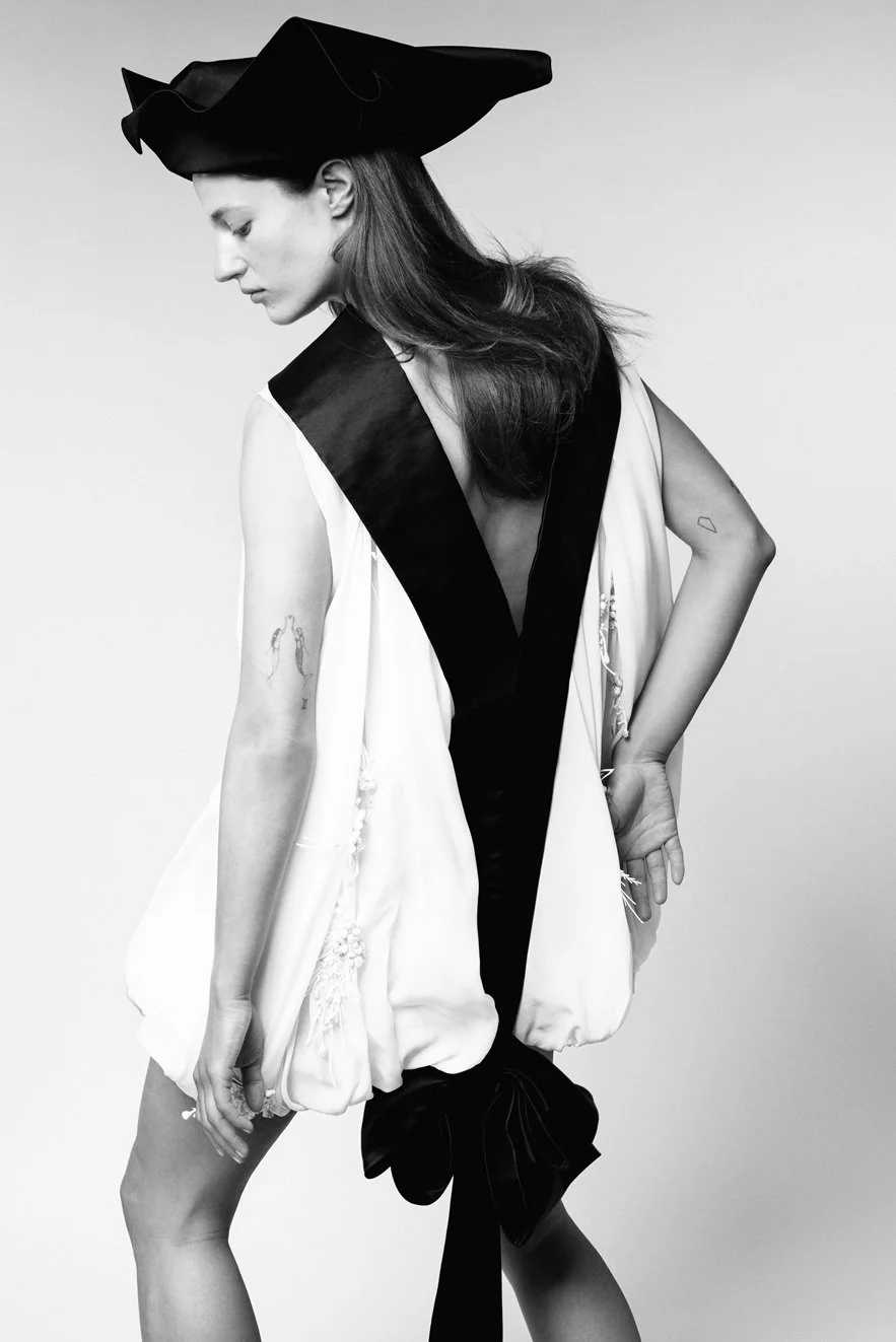 A black and white portrait of a woman wearing an oversized, structured hat and a flowing, sleeveless dress with contrasting dark and light sections. She has long hair, tattoos on her arms, and poses with her head tilted downward.