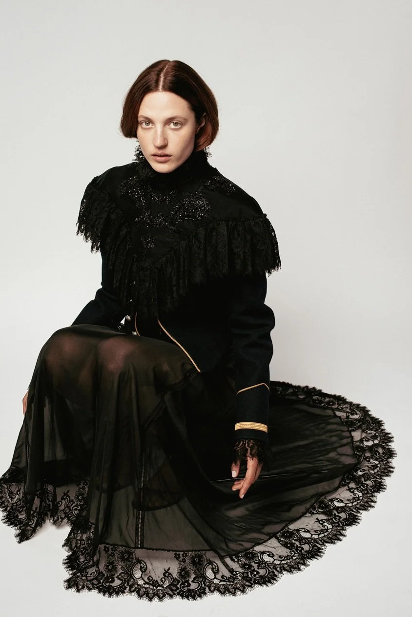 A woman with short red hair wearing a black lace and embroidered jacket and a sheer black lace skirt sitting on the floor against a plain gray background.