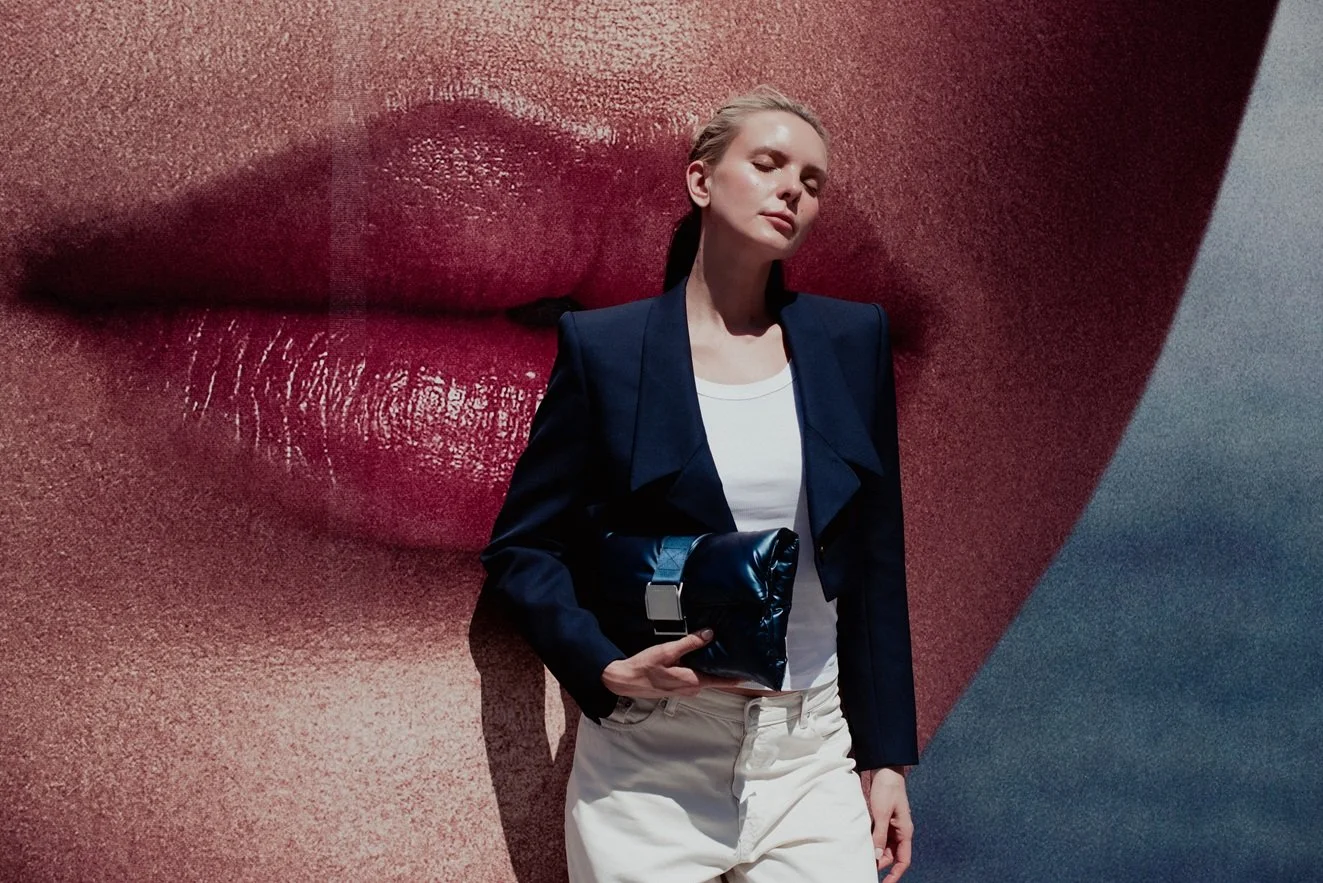 A woman stands in front of a large mural of lips on a wall, with her eyes closed and head slightly tilted. She is wearing a black blazer, a white shirt, and light-colored pants, and is holding a black clutch.