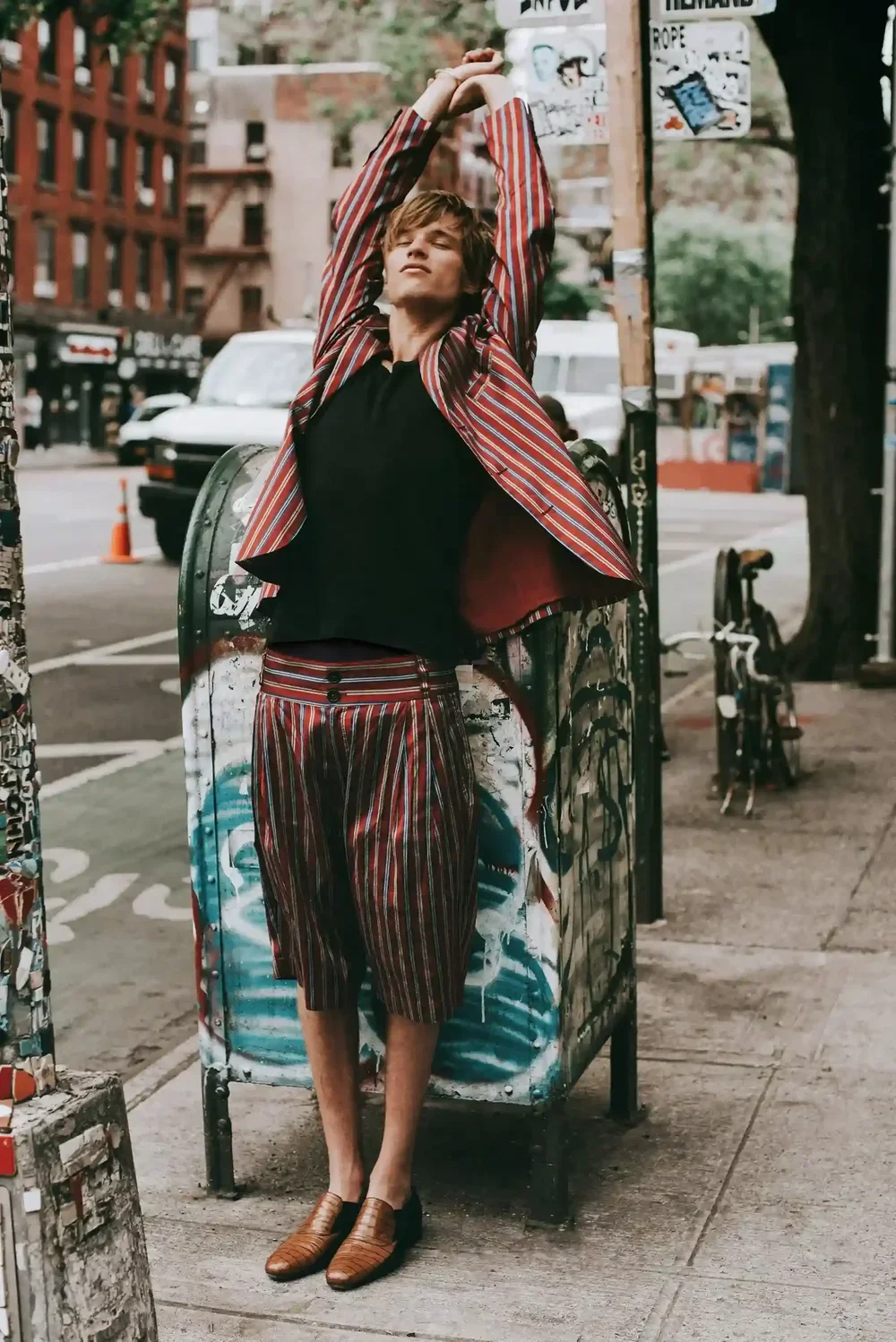 A young man in a colorful striped suit standing on a city sidewalk, stretching with arms above his head, near a graffiti-covered mailbox and a bike, with city buildings and parked cars in the background.