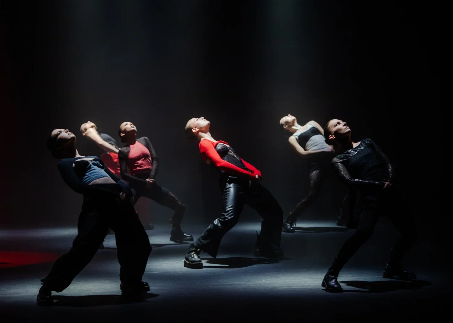 Six female dancers performing on stage with dramatic lighting, wearing colorful and black outfits, leaning back with their heads tilted upward.