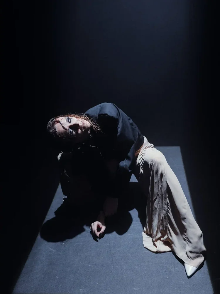 A person sitting on the floor in a dark room, illuminated by a single light source from above, wearing a dark top and beige wide-leg pants, with long hair and makeup.