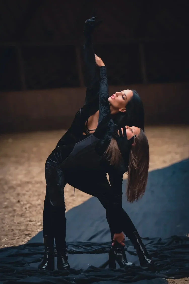 Two women dressed in black dance or pose together on dark fabric in an indoor setting, with one woman bent over and the other reaching above her in a dramatic pose.
