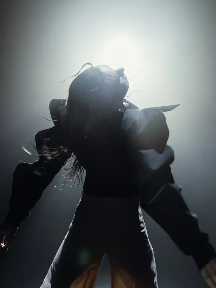 A person with long hair, wearing a hoodie and pants, is standing with their back to the camera, illuminated by a strong light from behind, creating a silhouette.