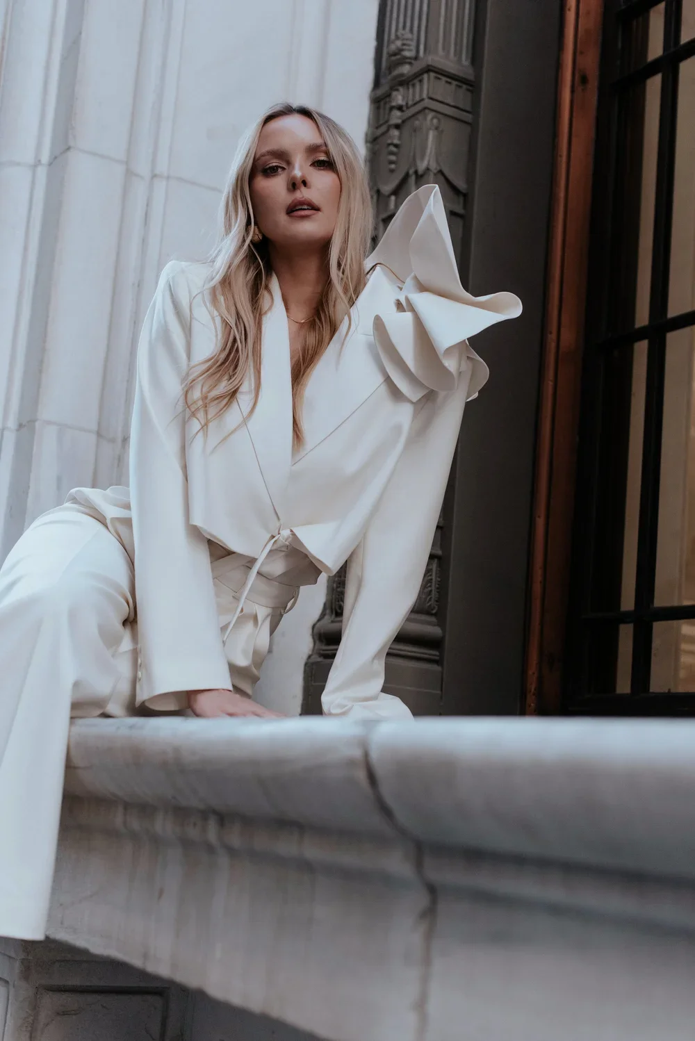 A woman with long blonde hair wearing an elegant white suit with ruffled shoulder details, sitting on a marble ledge in a historical building's interior with detailed architectural elements.