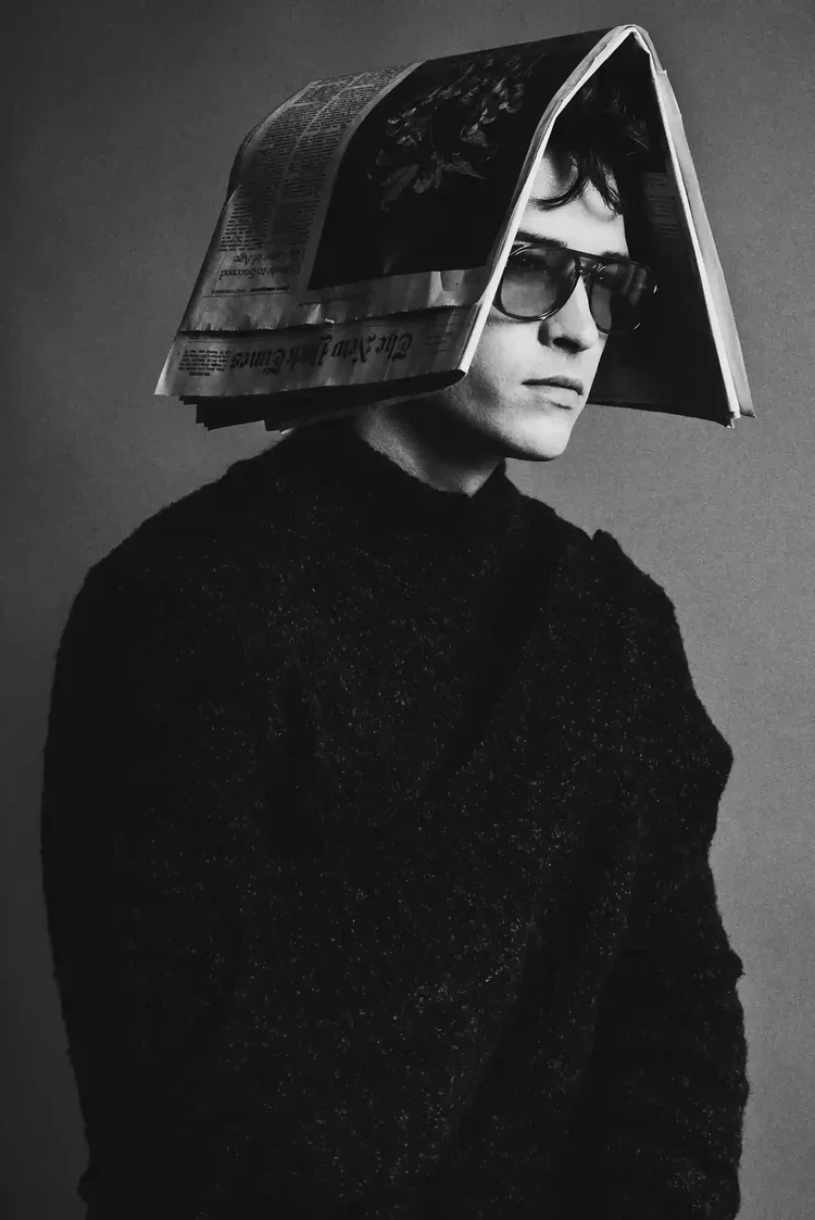 A black and white photo of a young man wearing glasses and a dark, textured sweater. A newspaper is draped over his head like a hat, partially covering his face.