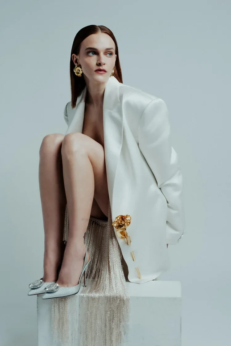 A woman with red hair and blue eyes, wearing large gold earrings, a white blazer, and high heels with embellishments, sitting on a white platform with her knees up and hands behind her back, against a plain light gray background.