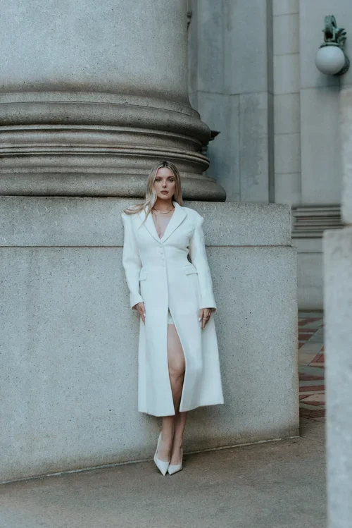 A woman in a white coat and high heels standing against a large stone column outside a building.
