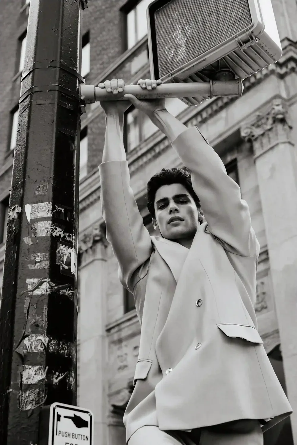 A young man in a light-colored suit is hanging from a streetlight pole with both hands during the daytime. The background shows an ornate building with classical architecture.