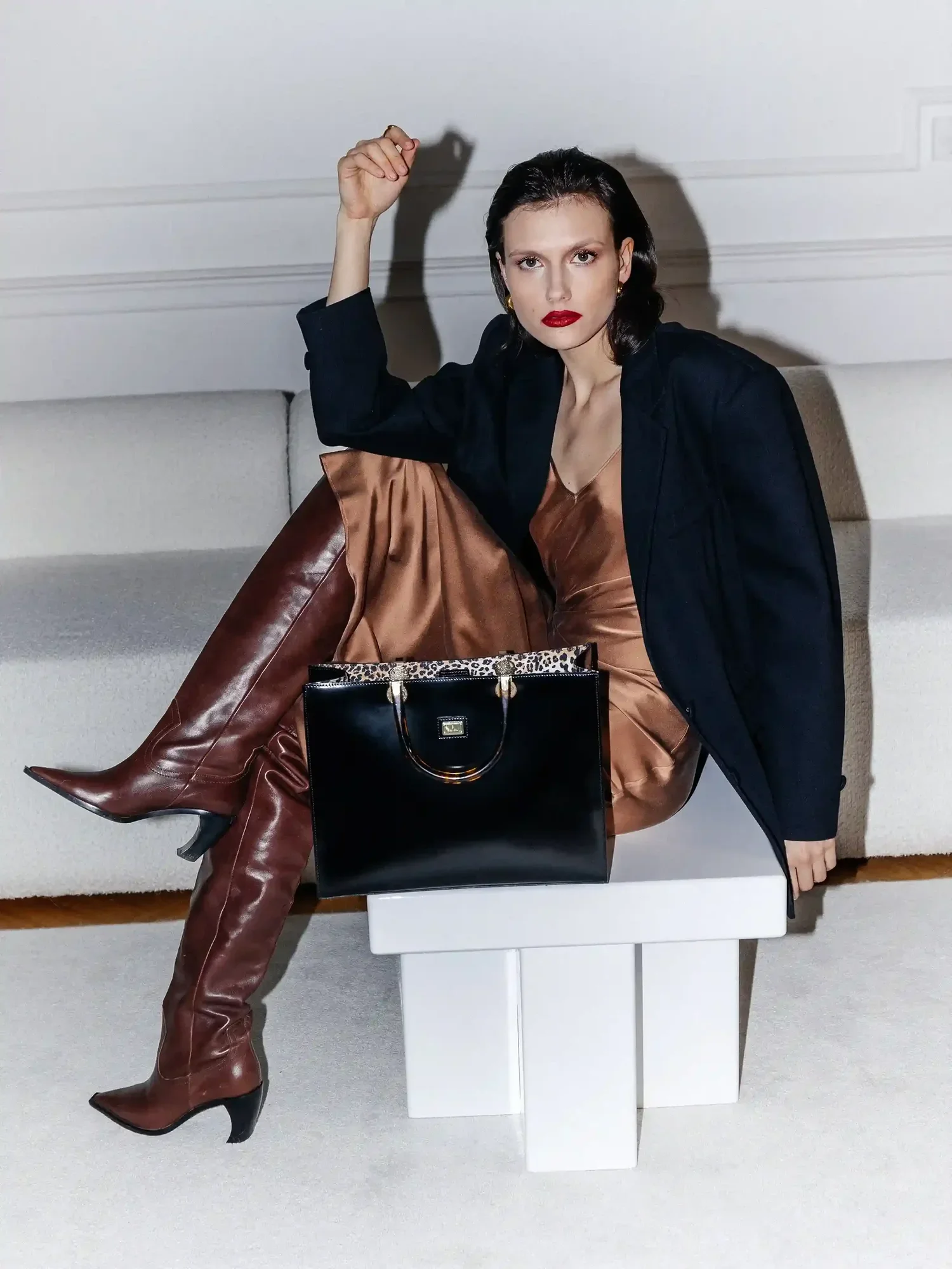 Woman sitting on a white block table, wearing a black blazer, brown leather pants, and brown high-heeled boots. She has dark hair, red lipstick, and is looking directly at the camera with a serious expression. A black handbag is placed on the table i