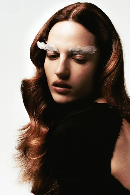 A woman with long, wavy brown hair with a reddish tint, wearing white feather eyebrows and a black top, looking down with a serious expression.