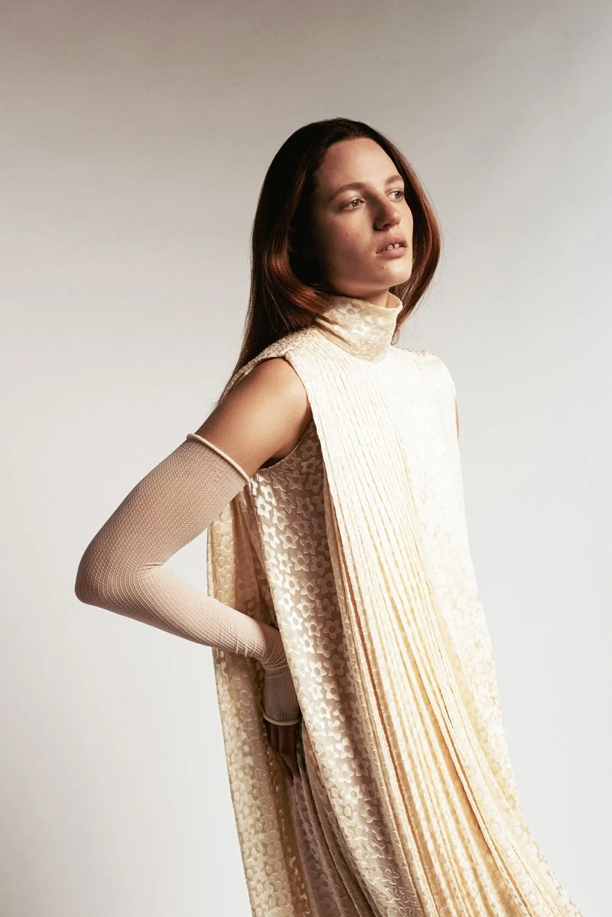 A woman with long reddish-brown hair posing in a sleeveless, beige, patterned dress with a high neck, paired with long beige gloves, against a plain light background.