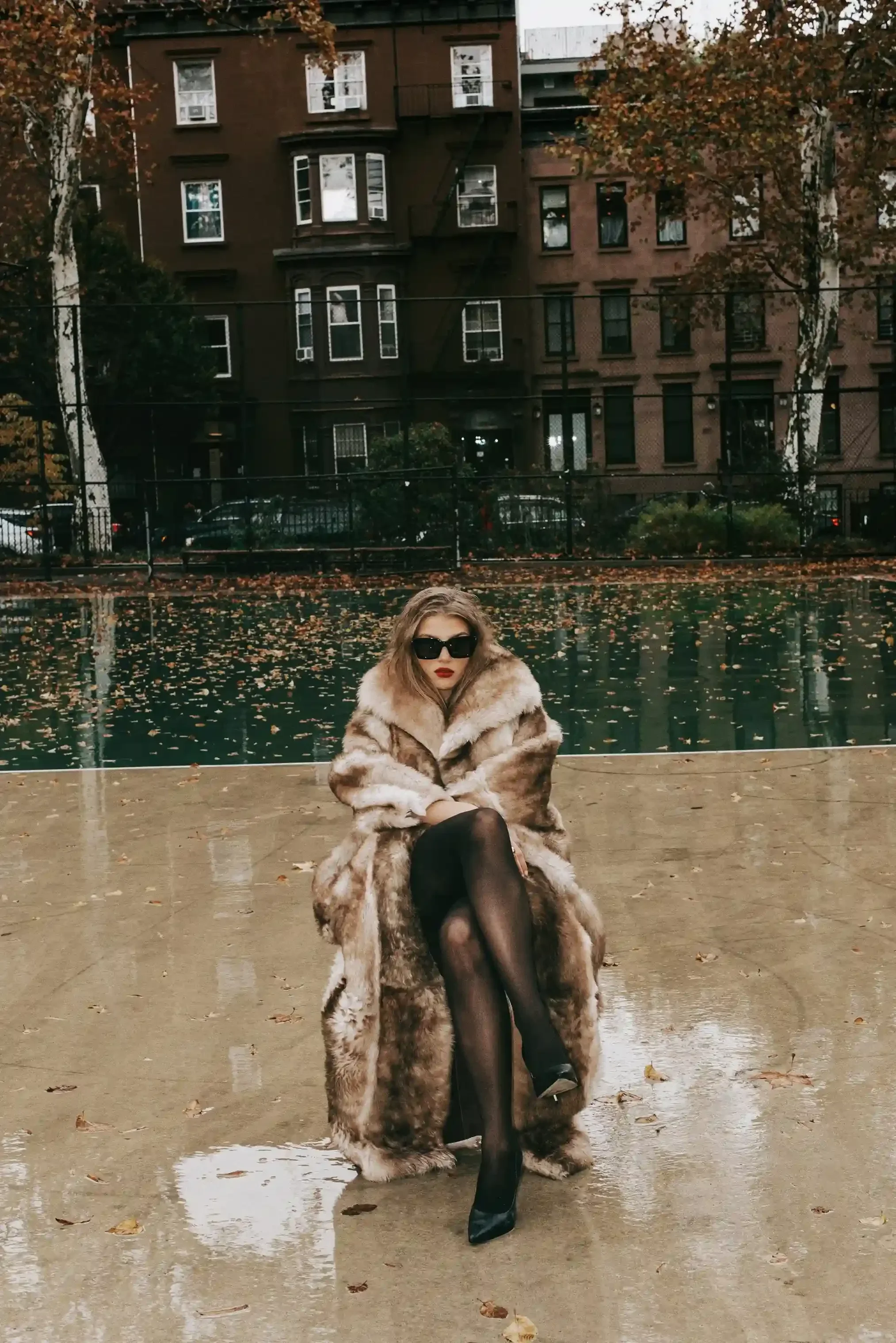 A woman sitting on the ground wearing black stockings, high heels, sunglasses, a fur coat, and red lipstick. She is outdoors near a body of water with fallen leaves, trees, buildings, and cars in the background.