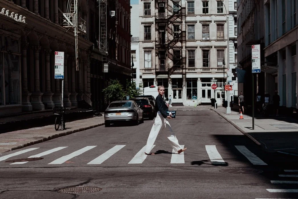 A woman crossing the street in an urban city setting, holding a camera or small device, with parked cars, tall buildings, and street signs visible.