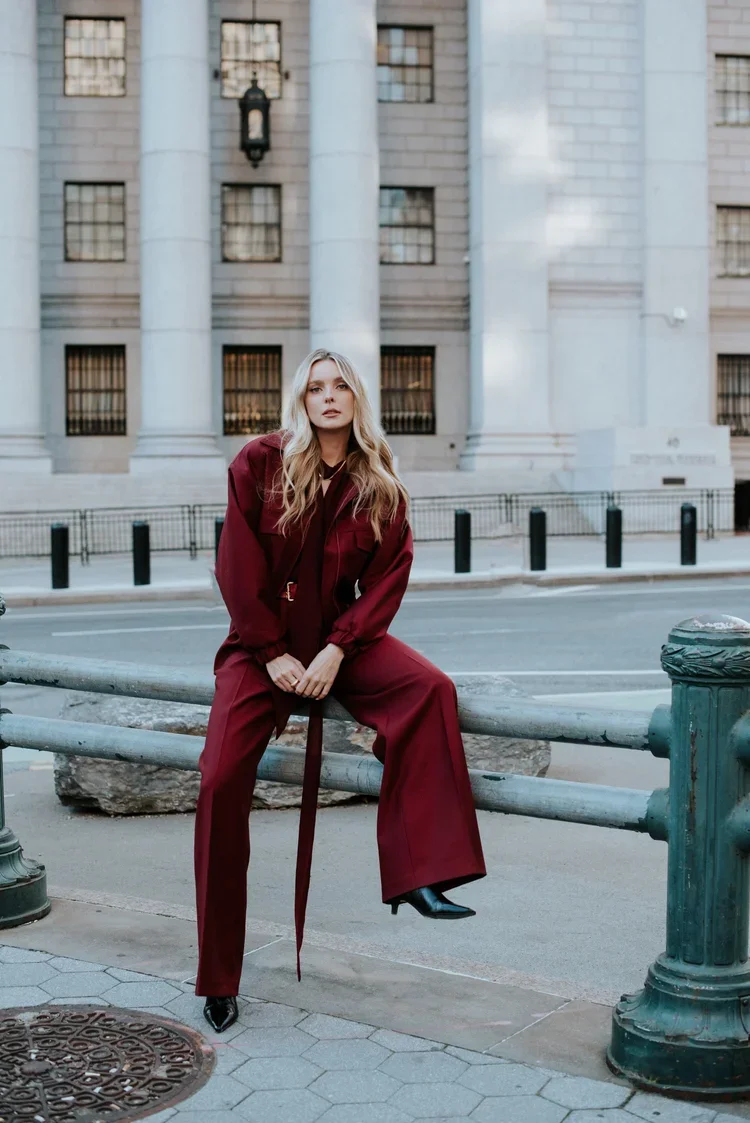 A young woman with long blonde hair sits on a metal barrier in front of a historic city building with large columns and windows, dressed in a burgundy outfit.