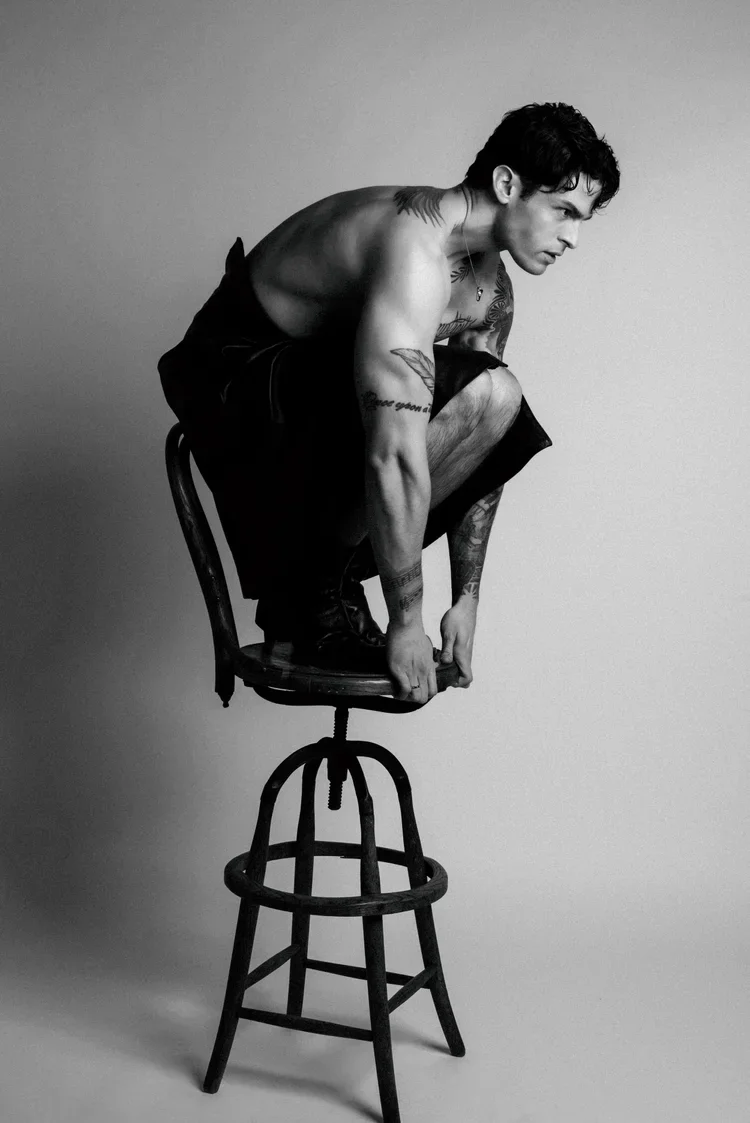 A black and white photo of a shirtless man with tattoos, squatting on top of a wooden chair with a tall seat, against a plain light background.