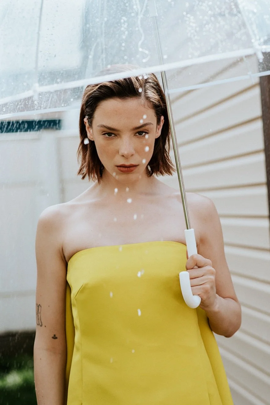 Young woman in a yellow strapless dress holding a clear umbrella with water droplets outside.