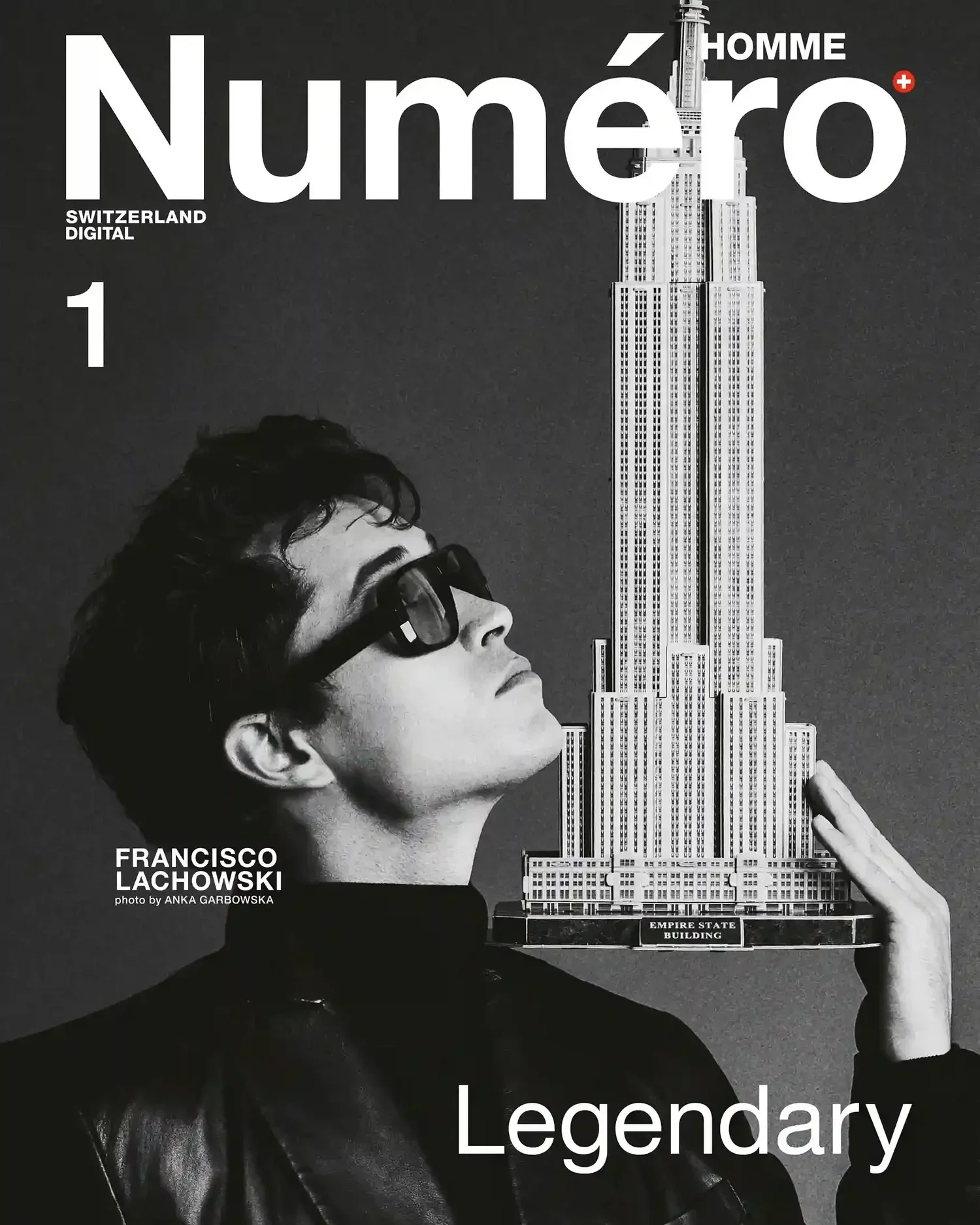 A black and white photograph of a woman with short hair and sunglasses, holding a miniature model of the Empire State Building. The text on the image indicates it is a cover of a magazine called 'Numéro Homme' from Switzerland. Additional text mentio