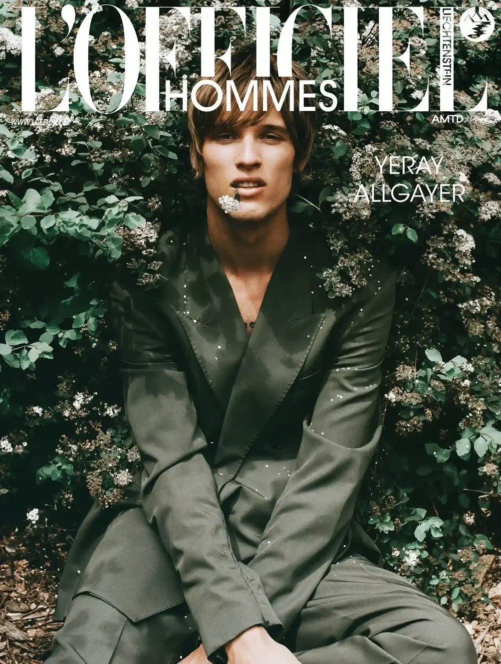 Cover of L'Officiel Hommes magazine featuring a man with medium-length hair, wearing a dark suit, sitting among green foliage and white flowers.
