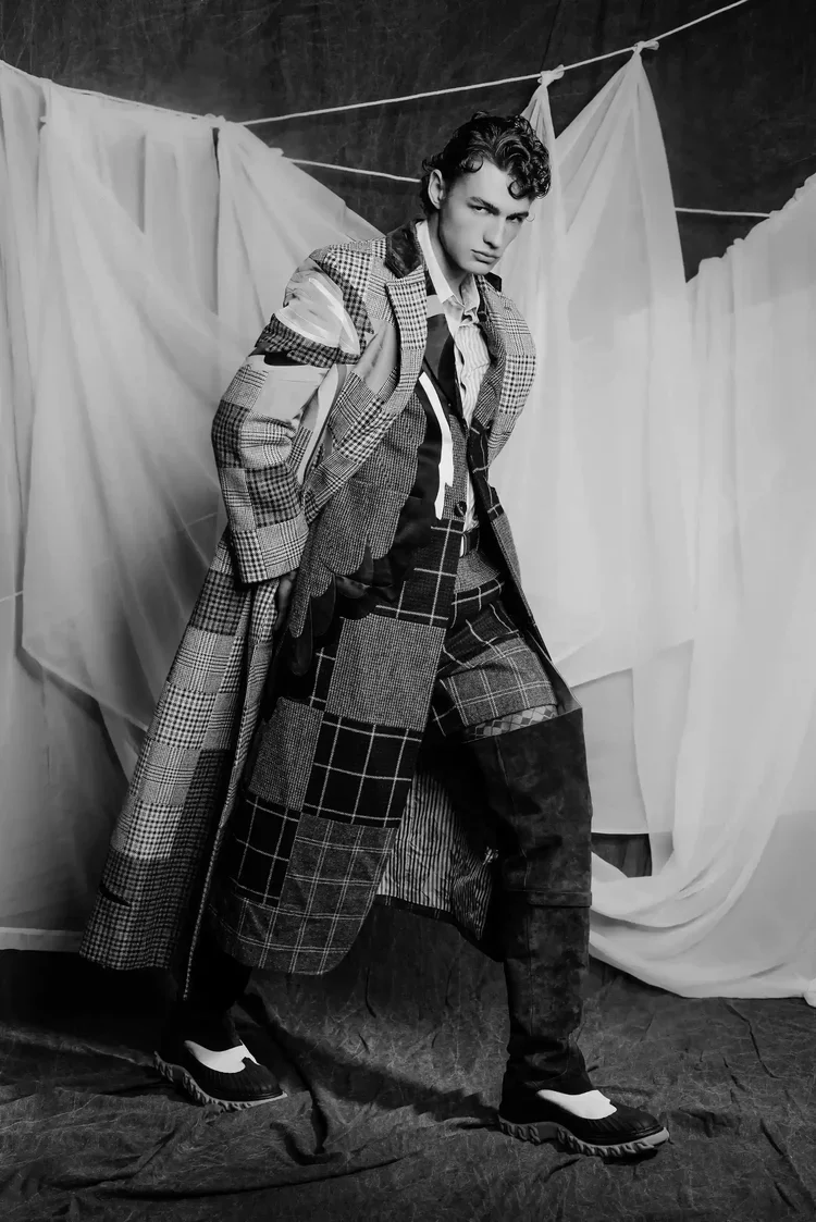 Black and white photo of a young man wearing a patchwork plaid coat, matching plaid pants, and hiking boots, standing in front of a backdrop of hanging white sheets.
