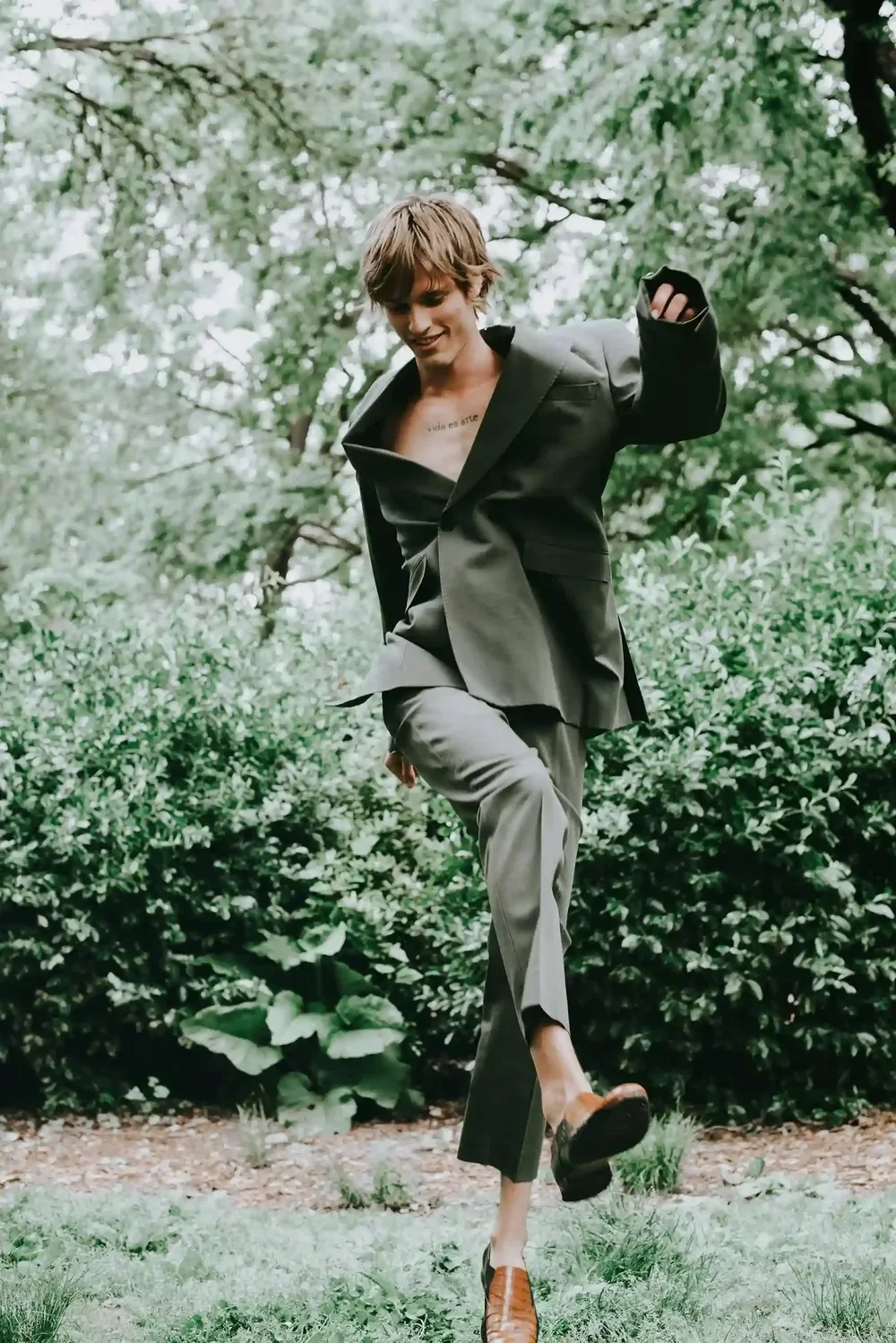 Young man with short brown hair, wearing a black blazer, gray pants, and brown shoes, balancing on one leg outdoors in a lush green garden.