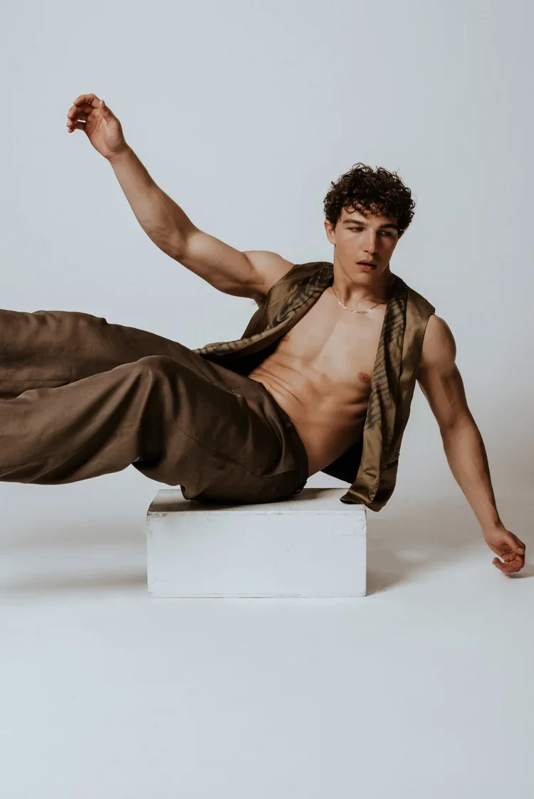 Young man with curly hair posing shirtless on a white block, wearing brown pants and a sleeveless open shirt, against a light gray background.
