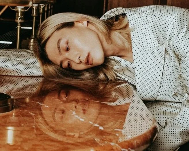 A woman with blonde hair lies on a wooden table, resting her head and cheek against a reflective surface, wearing a light-colored polka dot shirt.