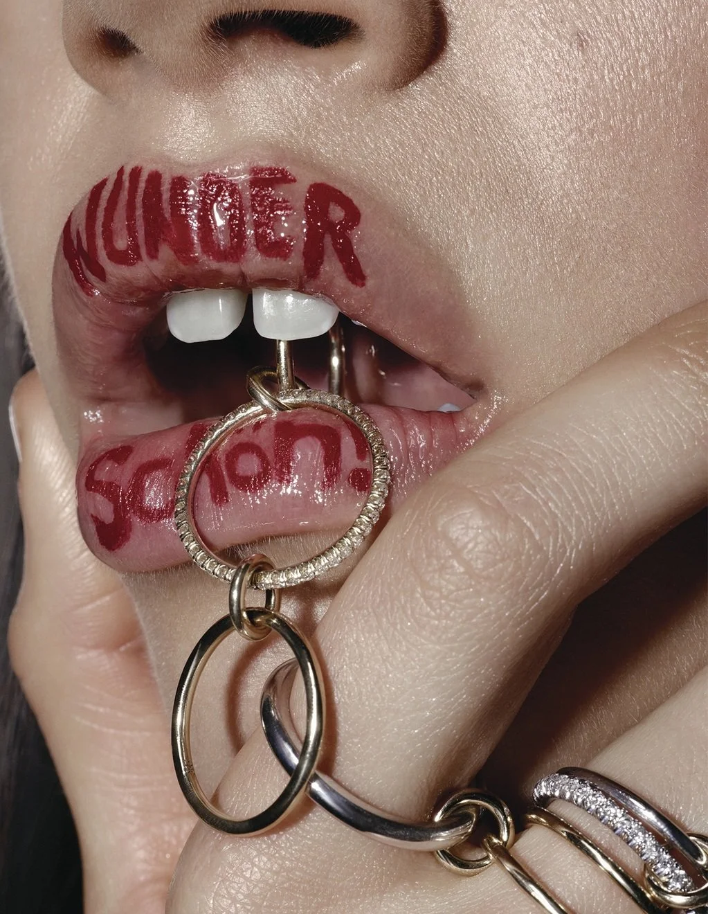 Close-up of a person's lips with red lipstick that reads 'HUNTER' and 'SCHOOL' with a tongue pierced by a hoop, and hand with rings touching lips.