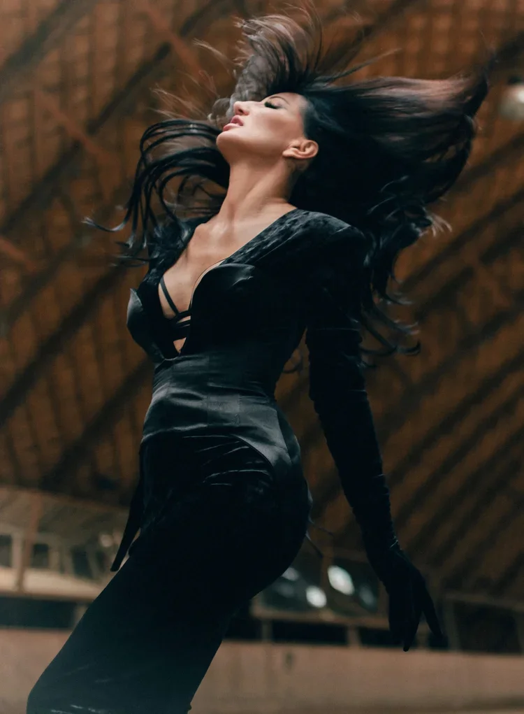 A woman with long black hair is dancing or spinning with her eyes closed inside a wooden building, wearing a black dress with cutouts.